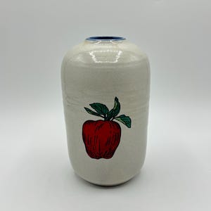 May include: A white ceramic vase with a blue rim and a red apple design.