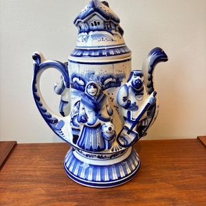 Könnte beinhalten: Blaue und weiße Keramik-Teekanne mit detaillierter Dorfszene. Die Teekanne hat einen Griff, einen Ausguss und einen hausförmigen Deckel. Das Design umfasst Figuren, einen Hund und eine Winterlandschaft. Die Teekanne steht auf einer Holzoberfläche.