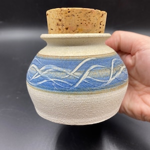 Op de afbeelding: Een kleine, witte keramische pot met een blauw en bruin ontwerp. De pot heeft een kurken deksel.