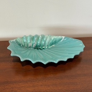 Puede incluir: Un plato de servir de cerámica de color verde azulado con forma de concha. El plato tiene un borde festoneado y un centro elevado y texturizado.