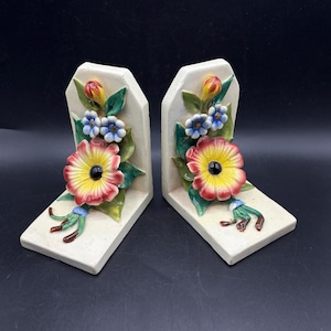 Puede incluir: Un par de sujetalibros de cerámica con un diseño floral. Los sujetalibros son blancos con un diseño en relieve de flores rojas, amarillas, verdes y azules.