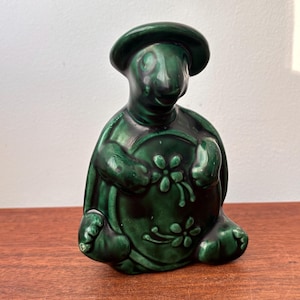 Op de afbeelding: Een donkergroen keramieken schildpadbeeldje met een hoed, dat een rond object met bloemmotieven vasthoudt. Het beeldje heeft een glanzende afwerking en staat op een houten oppervlak. De schildpad heeft gedetailleerde kenmerken.