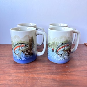 May include: Four ceramic coffee mugs with a fishing theme. Each mug features a colourful illustration of a trout leaping from the water, with a fisherman in the background. The mugs have a white background with a blue base and grey handle.