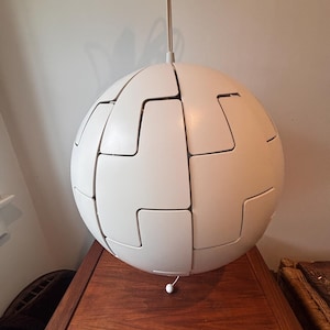 May include: A white, spherical pendant light. The light is made from interlocking, geometric shapes, forming a puzzle-like design. It hangs from a thin, off-white cord. The fixture is resting on a dark wooden surface.