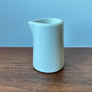 Op de afbeelding: Een kleine, lichtblauwe keramische roomkannetje met een gebogen handvat en een schenktuit. Het kannetje is cilindrisch met een gladde, glanzende afwerking. Het staat op een houten oppervlak tegen een lichtblauwe achtergrond.