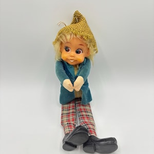 Puede incluir: Una muñeca vintage con un gorro de punto marrón, una chaqueta de pana azul, pantalones de cuadros rojos y blancos y zapatos negros. La muñeca tiene una cara pintada con cabello castaño y ojos grandes.