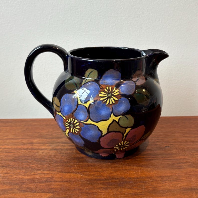 Op de afbeelding: Een donkerblauwe keramische kan met een handvat en tuit. De kan is versierd met grote blauwe, gele en kastanjebruine bloemmotieven. De kan staat op een houten oppervlak.