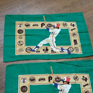 May include: Two green pillowcases with a baseball player in a white uniform swinging a bat. The pillowcases have a white border with a variety of baseball team logos, including the Pirates, Astros, Giants, Mets, and Royals.