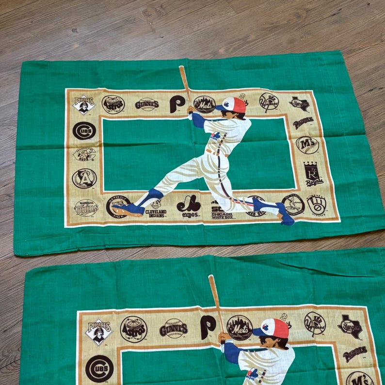 May include: Two green pillowcases with a baseball player in a white uniform swinging a bat. The pillowcases have a white border with a variety of baseball team logos, including the Pirates, Astros, Giants, Mets, and Royals.