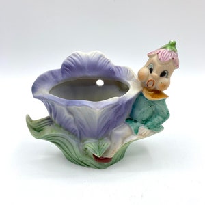 May include: A ceramic wall planter shaped like a purple flower with a green stem and leaves. A small, whimsical elf figure sits on the flower, with a pink flower on its head.