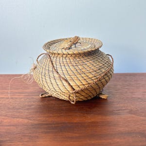 Può includere: Un cestino rotondo intrecciato a mano con coperchio. Il cestino presenta un motivo intrecciato nei toni del beige e del marrone. Un cordoncino marrone avvolge il cestino. Il coperchio è fissato con un piccolo pezzo di legno. Il cestino è su una superficie di legno.