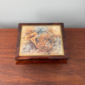May include: A square wooden music box with a detailed illustration of a French horn, angels, and musical notes. The box has a dark wood finish with a gold border around the artwork.