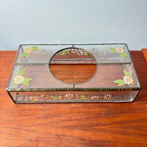 Puede incluir: Caja de pañuelos rectangular de vidrio con diseño floral. La caja presenta una parte superior y laterales de vidrio transparente, con margaritas blancas y hojas verdes pintadas. Una abertura circular está centrada en la parte superior.