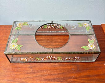 May include: A rectangular glass tissue box with a decorative floral design. The box features a clear glass top and sides, with painted white daisies and green leaves. A circular opening is centered on the top.
