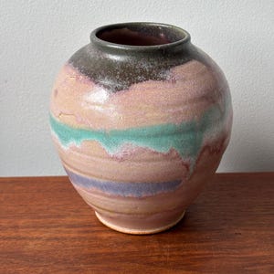 May include: A round ceramic vase with a wide body and a narrow opening. The surface features a multi-coloured glaze with horizontal bands of dusty pink, muted teal, and soft purple, topped with a dark brown rim and a textured, uneven finish.