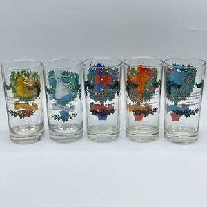 May include: Set of five clear glass tumblers with colorful illustrations and text. Each glass features a different design, including a partridge, a lady, drummers, a tower, and a swan. The glasses are decorated with festive greenery and text.