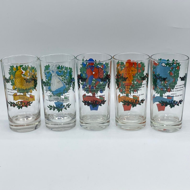 May include: Set of five clear glass tumblers with colorful illustrations and text. Each glass features a different design, including a partridge, a lady, drummers, a tower, and a swan. The glasses are decorated with festive greenery and text.