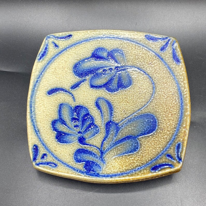 K&ouml;nnte beinhalten: Ein quadratischer Keramikteller mit einem blauen und wei&szlig;en Blumendesign. Der Teller hat eine hellbraune Glasur und einen leicht erh&ouml;hten Rand.
