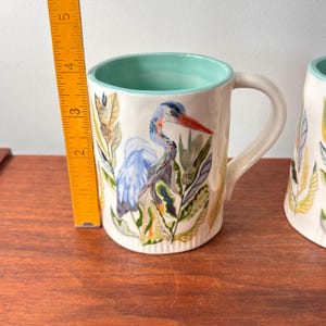 Puede incluir: Taza de cer&aacute;mica blanca con una ilustraci&oacute;n de garza azul, pico rojo y follaje verde y naranja. El interior de la taza es de color verde azulado claro. La taza mide aproximadamente 10 cm de alto.