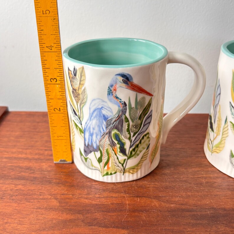 Puede incluir: Taza de cer&aacute;mica blanca con una ilustraci&oacute;n de garza azul, pico rojo y follaje verde y naranja. El interior de la taza es de color verde azulado claro. La taza mide aproximadamente 10 cm de alto.