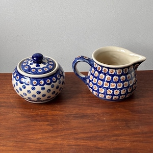 May include: A ceramic sugar bowl with a lid and a matching creamer jug. Both pieces feature a cream-coloured base with a repeating pattern of blue circles containing smaller brown dots. The sugar bowl has a round lid with a blue knob handle.
