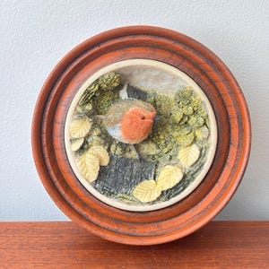 May include: A circular wall plaque featuring a three-dimensional robin with a bright orange breast, perched on a dark textured surface. The bird is surrounded by sculpted green foliage and pale yellow leaves, all encased within a polished wooden frame.