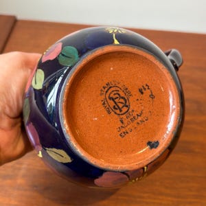 Op de afbeelding: Een donkerblauwe keramische kan met bloemmotieven in roze, groen en geel. De bodem van de kan is bruin en heeft de woorden "Royal Stanley Ware" en "Jacobian England" erop gedrukt.