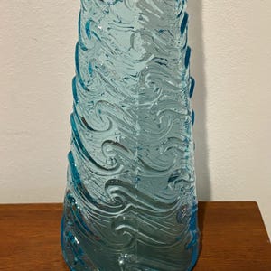 May include: A tall, clear blue glass vase with a swirling wave pattern. The vase is made of textured glass and has a wide base.