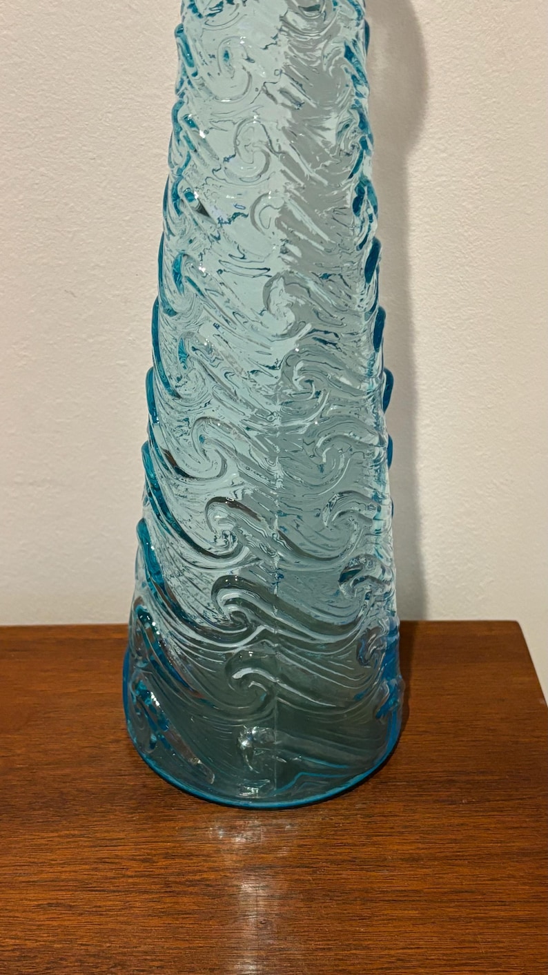 May include: A tall, clear blue glass vase with a swirling wave pattern. The vase is made of textured glass and has a wide base.