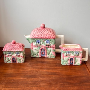 May include: A ceramic tea set with a whimsical cottage design. The set includes a teapot, sugar bowl, and creamer, all decorated with pink roofs, brick walls, and green ivy. The teapot has a handle and spout.