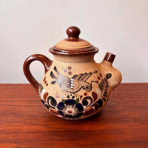 Op de afbeelding: Kleine keramische theepot met deksel, handvat en tuit. De theepot is beige met bruine accenten en is versierd met een handgeschilderd vogel- en bloemmotief in tinten blauw, bruin en groen. De theepot staat op een houten oppervlak.