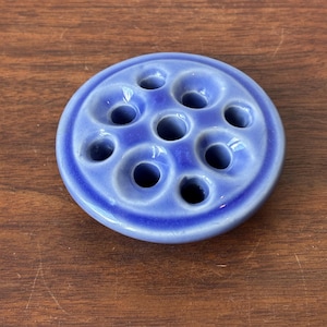 May include: A circular, periwinkle blue ceramic flower frog featuring a smooth, glazed finish. The object has a domed top surface with eight evenly spaced, circular holes arranged around a central ninth hole for holding flower stems upright.