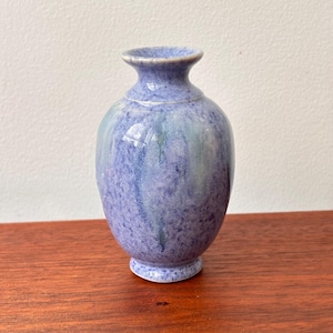 Puede incluir: Un pequeño jarrón de cerámica azul con un acabado moteado. El jarrón tiene un cuerpo redondeado, un cuello estrecho y un borde acampanado. El color es una mezcla de azules claros y oscuros con toques de verde, creando un aspecto artístico único. El jarrón se asienta sobre una pequeña base circular.