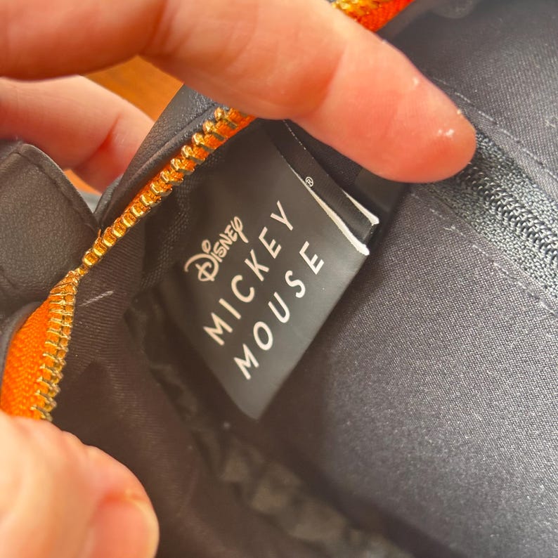 May include: Close-up of a black bag with a gold zipper and orange trim. The tag reads "Disney MICKEY MOUSE" in white lettering. The bag appears to be made of a durable material.