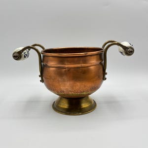 May include: A copper planter with two white ceramic handles. The planter has a hammered finish and sits on a brass base.