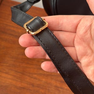 May include: Close-up of a black leather strap with a gold-colored metal buckle. The strap is stitched with a contrasting thread. The background is a blurred wooden surface.