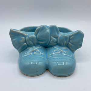 Könnte beinhalten: Ein hellblauer Keramik-Pflanztopf in Form von Schuhen mit einer Schleife oben.