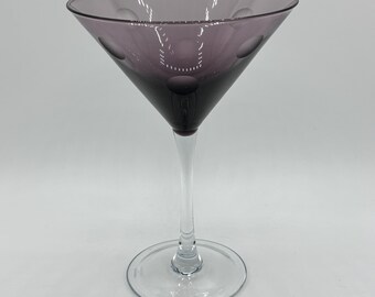 May include: A clear glass martini glass with a purple tint and a textured design. The glass has a long stem and a wide, shallow bowl.