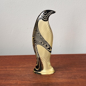 May include: A decorative penguin figurine with a gold body and black, wave-like patterns. The penguin has a curved neck and a small base. The figurine is made of glass or a similar material and is displayed on a wooden surface.
