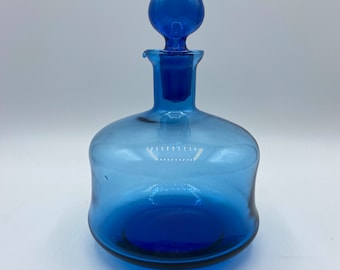 May include: A cobalt blue glass decanter with a round, bulbous stopper. The decanter has a wide, round base and a slightly flared neck.