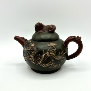 Op de afbeelding: Een klein groen theepotje met een drakenontwerp en een houten deksel. De draak is goudkleurig en het theepotje heeft een houten handvat.