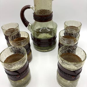 Puede incluir: Un juego de siete vasos transparentes de vidrio y una jarra con bandas de cuero marrón. Los vasos y la jarra tienen una textura de vidrio craquelado.
