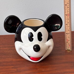 May include: A ceramic Mickey Mouse head-shaped planter. The planter features black ears, a black nose, and a red mouth. The interior is beige. The planter is on a wooden surface.
