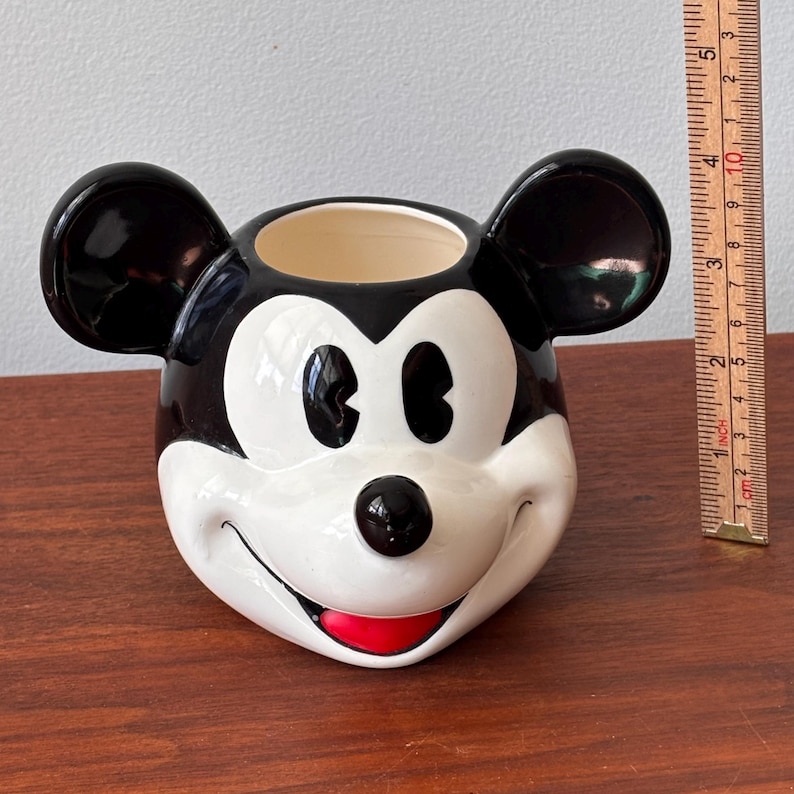 May include: A ceramic Mickey Mouse head-shaped planter. The planter features black ears, a black nose, and a red mouth. The interior is beige. The planter is on a wooden surface.