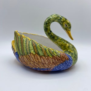 Peut inclure: Un pot de fleurs en céramique en forme de cygne avec un motif peint vert, bleu et marron. Le cygne a un bec jaune et des yeux noirs.