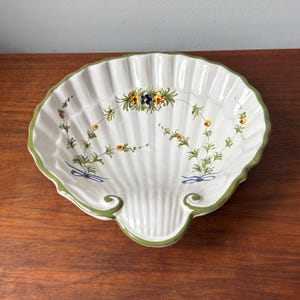 May include: A white ceramic serving dish shaped like a large scallop shell with a green painted rim. The interior features hand-painted floral motifs in yellow, blue, and green, with delicate stems and small blue bows at the base of the design.