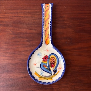Puede incluir: Un reposacucharas de cerámica con un diseño azul y blanco. El reposacucharas presenta una ilustración de gallo en azul, rojo y amarillo. El mango tiene un diseño trenzado amarillo y naranja con un borde azul. El reposacucharas mide unos 25 cm de largo.