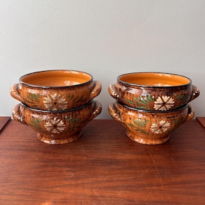 May include: Four ceramic soup bowls with handles, stacked in pairs. The bowls are brown with orange interiors and decorated with white floral and green leaf designs. The bowls are on a wooden surface.