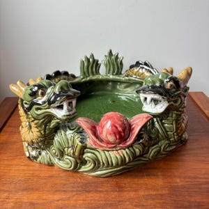 May include: A circular ceramic bowl featuring two sculpted dragon heads facing each other. The dragons are primarily green with black accents and tan horns. A central red, lotus-like flower ornament sits between the dragon heads. The bowl has a glossy finish.