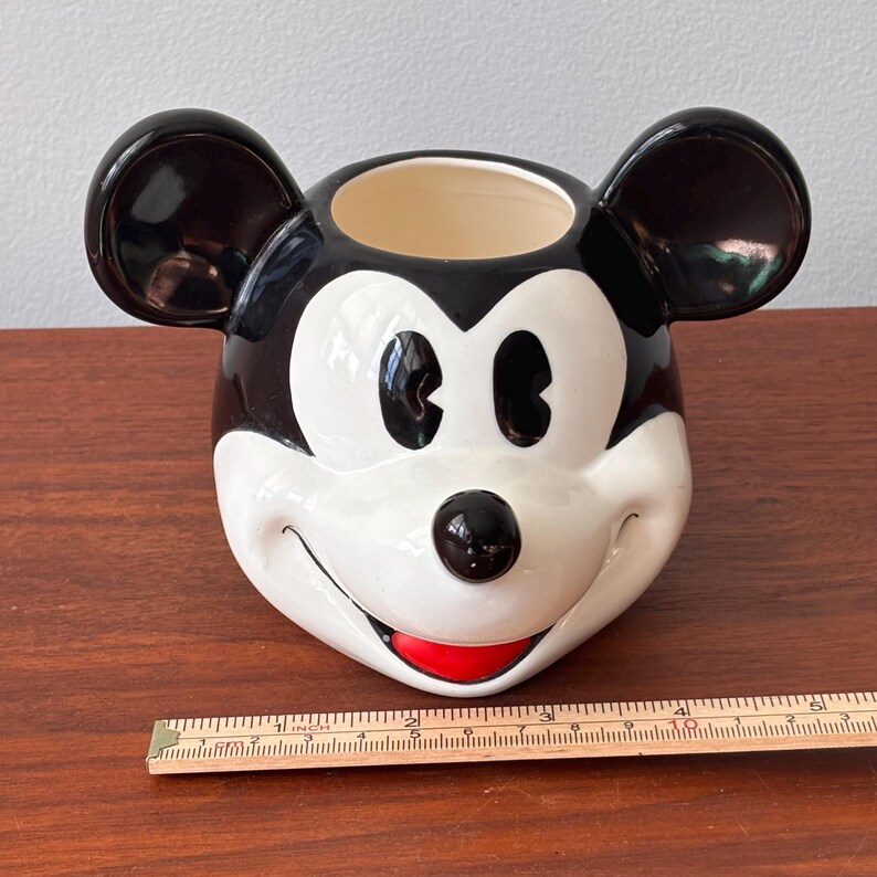 May include: A ceramic Mickey Mouse head-shaped planter. The planter is primarily white with black ears and a black top. It features a black nose, black eyes, and a red smile. A ruler is placed next to the planter.